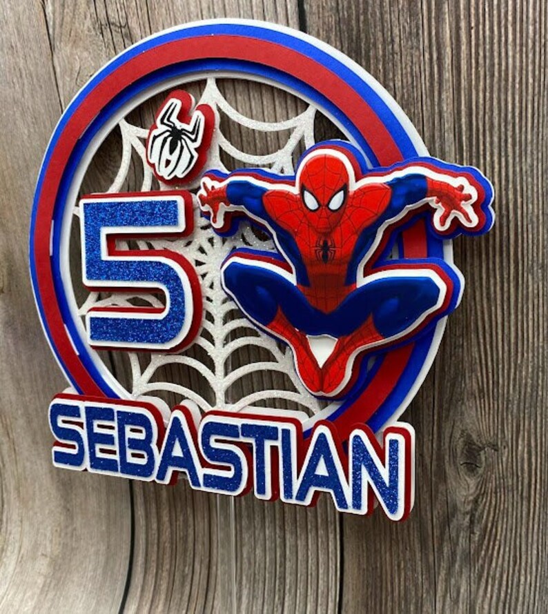 May include: A red, blue and white cake topper featuring Spiderman, a spiderweb and the number 5. The name Sebastian is written in blue glitter letters.