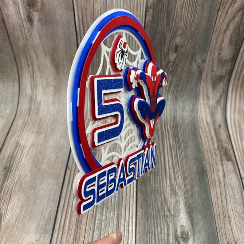May include: A red, white and blue cake topper featuring a Spiderman figure, a web design and the number 5. The topper also includes the name "Sebastian".