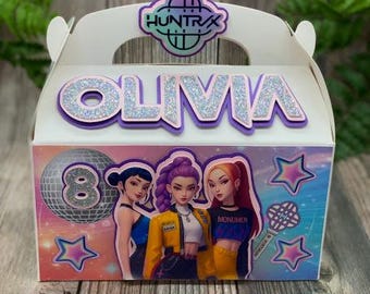 Peut inclure: Une boîte à cadeaux blanche avec une poignée, avec le nom "OLIVIA" en lettres pailletées. La boîte est décorée d'illustrations de trois filles, d'étoiles, d'une boule disco et du texte "HUNTRAX".