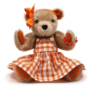May include: A plush teddy bear with light brown fur, wearing an orange and white checkered dress and a matching bow. The bear has orange accents on its ears and paws, and a red beaded bracelet.