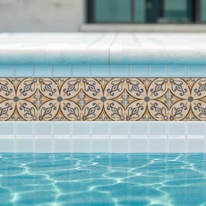 Puede incluir: Primer plano de una piscina con un borde de azulejos decorativos. El borde presenta un patrón repetitivo de diseños florales beige, marrones y azules. El agua de la piscina es de un color azul claro y transparente, con reflejos de sol.