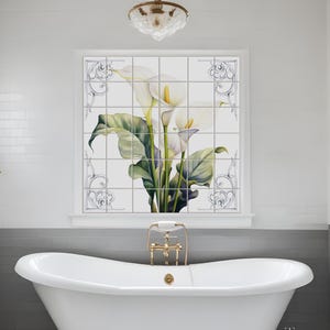 Puede incluir: Lirios blancos con hojas verdes se representan en un salpicadero de azulejos blancos en un baño. El salpicadero está sobre una bañera blanca con grifería dorada. El baño tiene paredes de azulejos blancos tipo metro y una pared de acento gris.