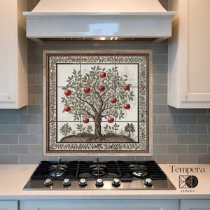 Op de afbeelding: Een witte keukenachterwand met een handgeschilderde keramische tegelmuur met een afbeelding van een rode appelboom. De tegelmuur is omlijst met een decoratieve rand. De achterwand bevindt zich boven een roestvrijstalen fornuis met zwarte knoppen.