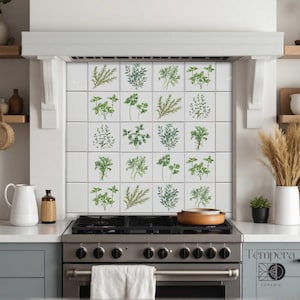 May include: White ceramic tile backsplash featuring watercolor illustrations of various herbs. The tiles are arranged in a grid pattern, showcasing green and gray-green botanical designs. The kitchen setting includes a stainless steel stove and white cabinetry.