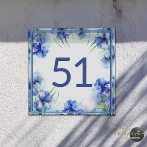 Puede incluir: Un letrero de número de casa de cerámica blanca con flores de iris azules de acuarela y el número 51 en azul.