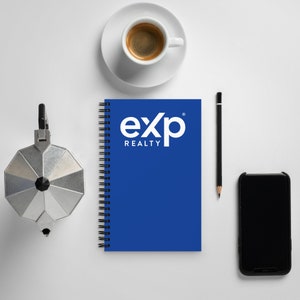 Puede incluir: Una libreta azul de espiral con el texto "exp realty" en blanco. Una cafetera de plata, una taza de café, un lápiz y un teléfono inteligente negro también están sobre la superficie blanca.