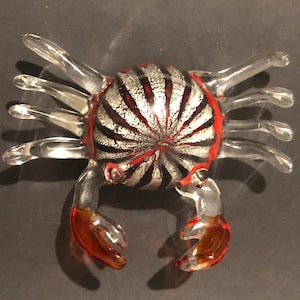 May include: A glass crab figurine with clear legs and claws. The crab's body is silver and red with a radial pattern. The claws are a reddish-orange colour. This decorative item is suitable for coastal or nautical themed decor.