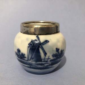 May include: A small, round, white ceramic jar with a silver rim. The jar is decorated with a blue and white windmill scene.