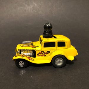 May include: A yellow toy car with a black top and flame decals. The car has a detailed engine and exhaust pipes. The wheels are black and the car is approximately 10 cm long.