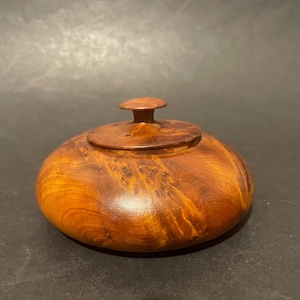 May include: A small, round, polished wooden lidded container with a smooth, glossy finish. The wood displays a rich, warm amber colour with intricate, swirling grain patterns and natural burl markings across the surface and the lid.