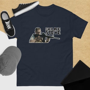 May include: A navy blue t-shirt with a white graphic of a man holding a rifle. The graphic includes the text "Be Like Steve".