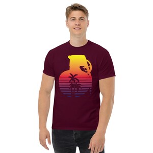 May include: A maroon t-shirt with a graphic of a yellow grenade silhouette with palm trees inside. The grenade is set against a sunset gradient background with orange, yellow, red, and purple stripes.