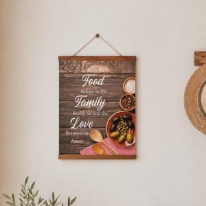 May include: A rectangular wooden wall hanging featuring a dark wood grain background with the text Bless the Food before us the Family beside us and the Love between us Amen. It includes a bowl of olives, a small bowl of spices, and two wooden spoons.