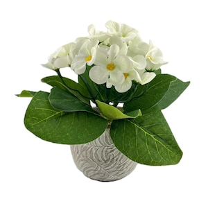 May include: A white artificial flower arrangement in a gray planter. The flowers have yellow centers and are arranged in a cluster. The leaves are green and lush.