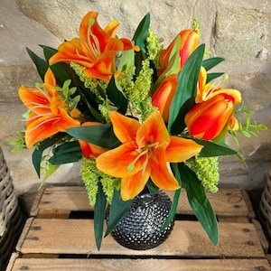 以下が含まれることがあります： オレンジ色のユリとチューリップ、緑の葉をあしらった鮮やかなフラワーアレンジメント。テクスチャ加工された濃いグレーの花瓶に入っています。木製の木枠の上に置かれ、素朴な雰囲気を醸し出しています。