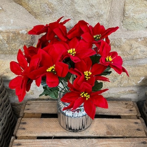 May include: A bouquet of vibrant red poinsettia flowers with yellow centers in a silver mercury glass vase. The arrangement sits on a rustic wooden crate, perfect for holiday decor. The flowers have a velvety texture.