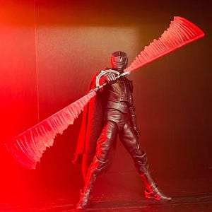 May include: A black action figure of a character in a dark outfit, cape, and helmet, holding a large, double-bladed red lightsaber. The figure is posed against a dark background with red lighting.