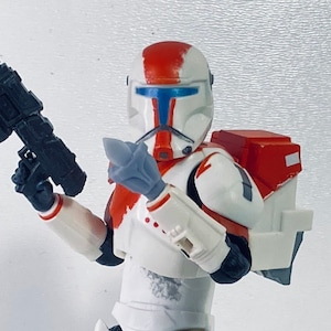 Puede incluir: Una figura de acción blanca y roja de un soldado clon de Star Wars con un rifle bláster negro. La figura tiene una pintura desgastada y sostiene un rifle bláster en su mano derecha.