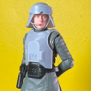 May include: A Star Wars Imperial Officer action figure. The figure is wearing a gray uniform, a chest plate, and a helmet. The figure is also wearing black boots and has a black holster. The background is yellow.