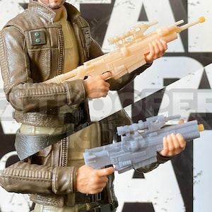 Könnte beinhalten: Nahaufnahme einer Spielzeug-Actionfigur, die zwei verschiedene Spielzeug-Blaster hält. Die Blaster sind beide grau und haben ein Zielfernrohr oben drauf. Die Figur trägt eine braune Jacke und einen schwarzen Gürtel.