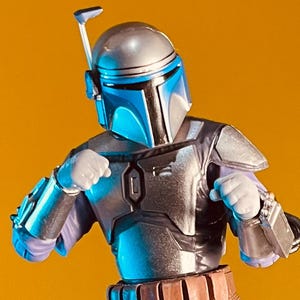 Puede incluir: Figura de acción detallada de un personaje con casco y armadura plateados y azules, con cinturón y pistoleras marrones. La figura posa sobre un fondo amarillo.