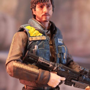 May include: A detailed action figure of a man with brown hair and a mustache, wearing a blue vest with yellow accents over a brown jacket. The figure is holding a black rifle. The figure is from the Star Wars franchise.
