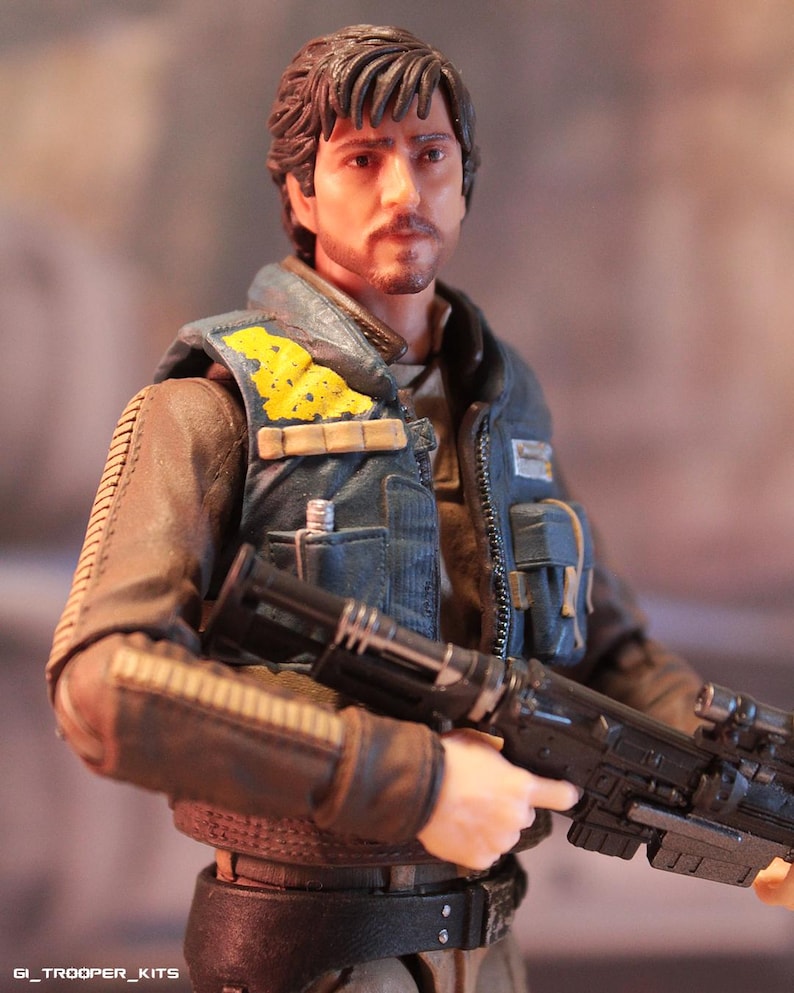 May include: A detailed action figure of a man with brown hair and a mustache, wearing a blue vest with yellow accents over a brown jacket. The figure is holding a black rifle. The figure is from the Star Wars franchise.
