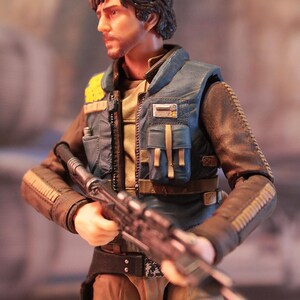 May include: Action figure of a character in a blue vest and brown jacket, holding a rifle. The figure has a serious expression and short, dark hair. The vest has yellow accents and multiple pockets. The figure is wearing a belt and has a holster.