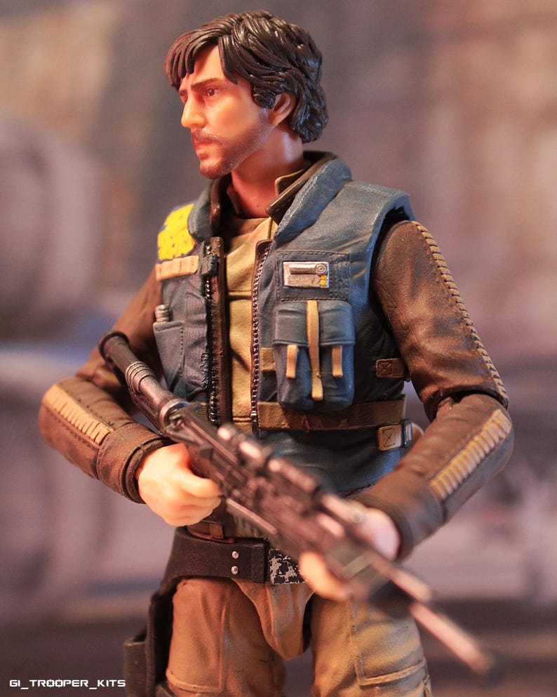 May include: Action figure of a character in a blue vest and brown jacket, holding a rifle. The figure has a serious expression and short, dark hair. The vest has yellow accents and multiple pockets. The figure is wearing a belt and has a holster.