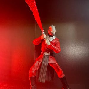 May include: A red action figure of a character in a dynamic pose, holding a large, serrated red blade. The figure is clad in a red uniform with black boots and a silver helmet. The background is dark, with red lighting.