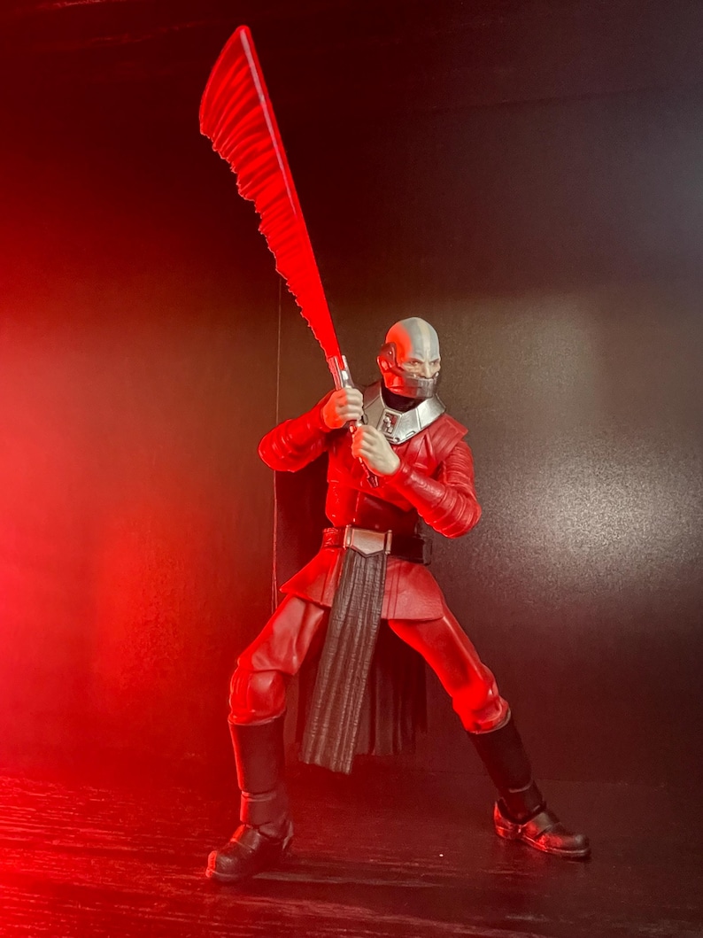 May include: A red action figure of a character in a dynamic pose, holding a large, serrated red blade. The figure is clad in a red uniform with black boots and a silver helmet. The background is dark, with red lighting.