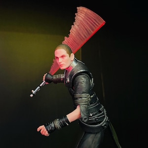 May include: A detailed action figure of a character in a black outfit, holding a red lightsaber. The figure has a stern expression and red eyes. The lightsaber has a unique, jagged blade design.