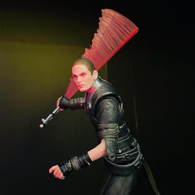 May include: A detailed action figure of a character in a black outfit, holding a red lightsaber. The figure has a stern expression and red eyes. The lightsaber has a unique, jagged blade design.