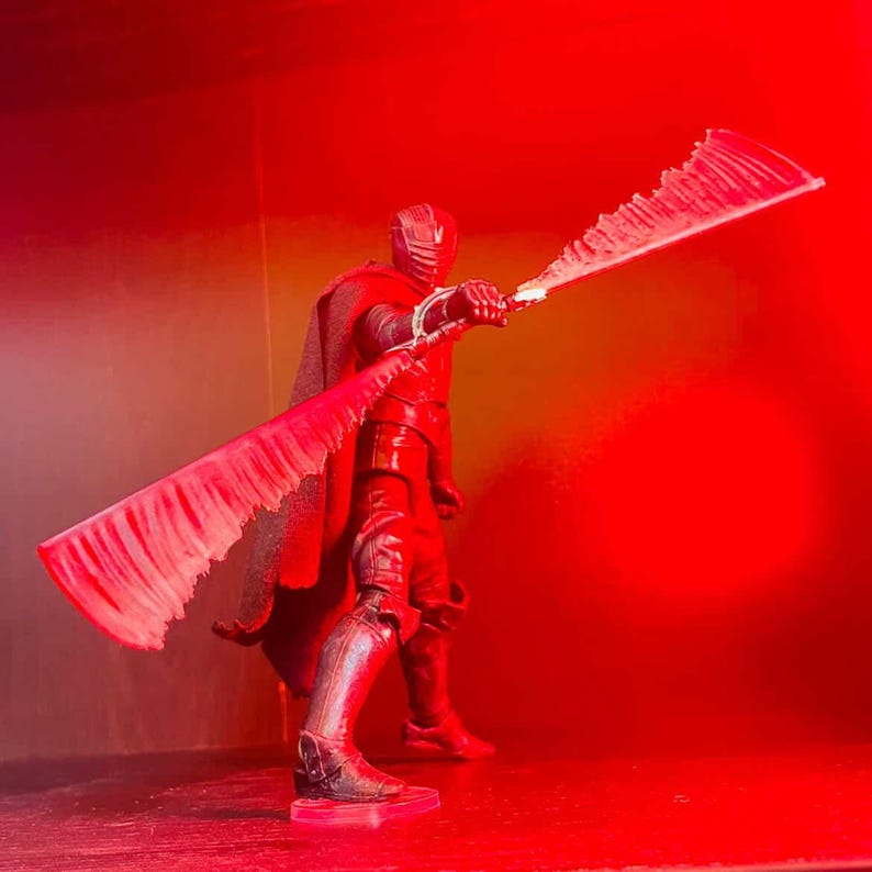 May include: A red action figure of a character in a black suit and cape, holding a large, serrated, red blade. The figure is posed against a red background, illuminated by red lighting.