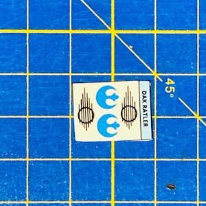 May include: A white sticker with two blue Rebel Alliance symbols and brown lines. The text "DAK RATTLER" is printed vertically on the right side of the sticker.