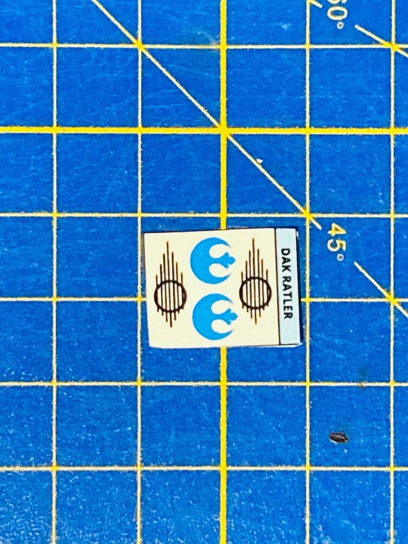 May include: A white sticker with two blue Rebel Alliance symbols and brown lines. The text "DAK RATTLER" is printed vertically on the right side of the sticker.