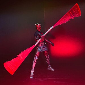 May include: A detailed action figure of Darth Maul, a character from Star Wars, holding a double-bladed lightsaber. The figure is in a dynamic pose, with a red and black color scheme. The lightsaber blades are a vibrant red.