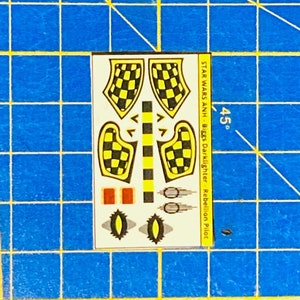 May include: A sheet of decals for a Star Wars model kit. The decals feature black and yellow checkered patterns, red rectangles, and black and white circles. The text on the sheet reads "STAR WARS ANH - Biggs Darklighter - Rebellion Pilot".