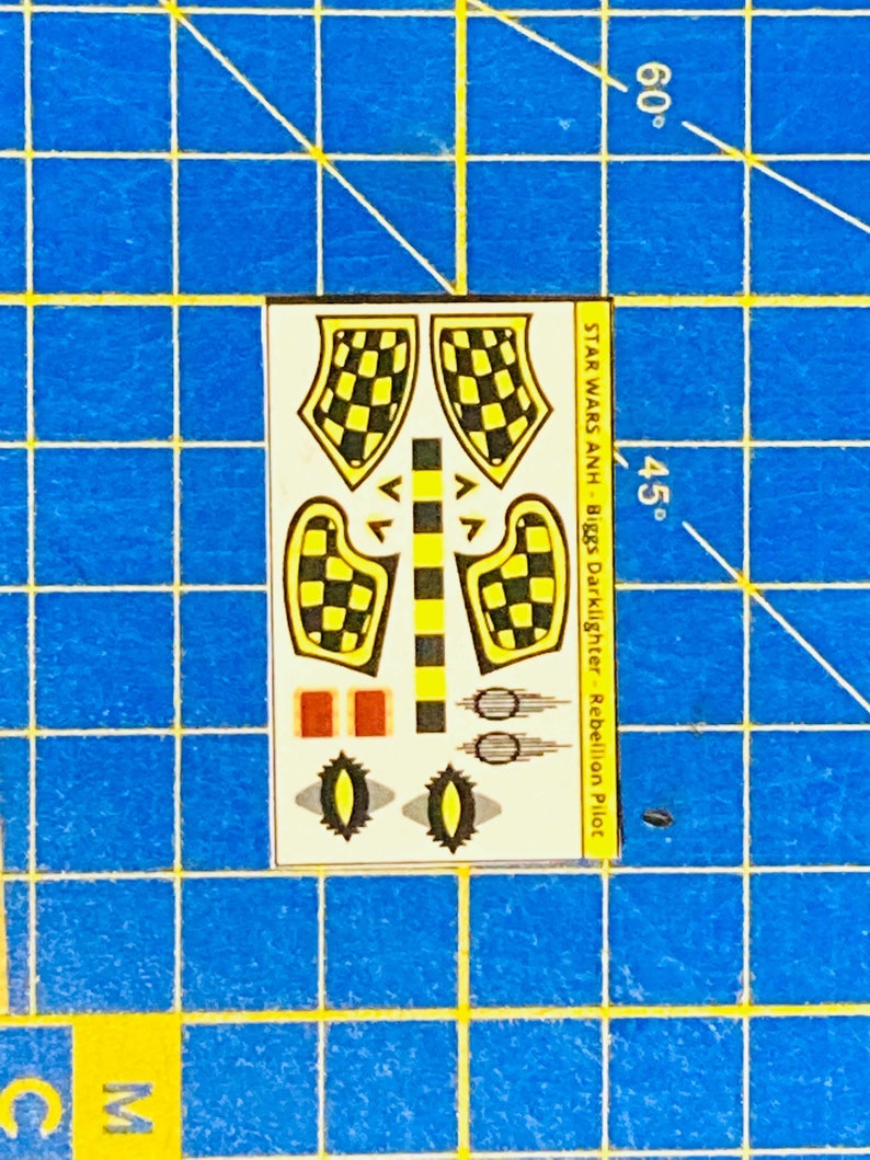 May include: A sheet of decals for a Star Wars model kit. The decals feature black and yellow checkered patterns, red rectangles, and black and white circles. The text on the sheet reads "STAR WARS ANH - Biggs Darklighter - Rebellion Pilot".