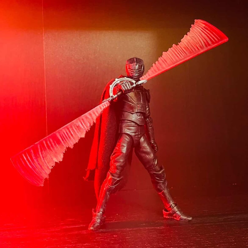 May include: A black action figure of a character in a dark outfit, cape, and helmet, holding a large, double-bladed red lightsaber. The figure is posed against a dark background with red lighting.