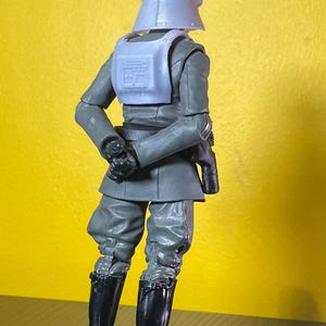 May include: A gray action figure of a character in a military uniform, with a helmet, vest, and black boots. The figure is posed with its back to the viewer, set against a yellow background.