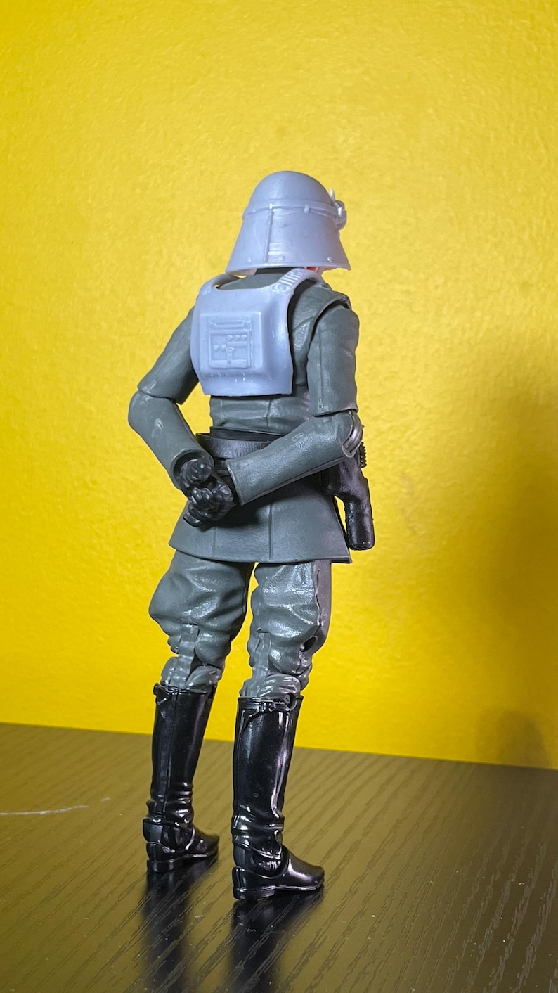 May include: A gray action figure of a character in a military uniform, with a helmet, vest, and black boots. The figure is posed with its back to the viewer, set against a yellow background.