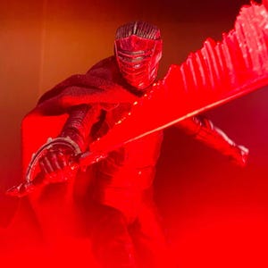 May include: A red action figure of a character in a black suit and helmet, holding a large red lightsaber. The figure is posed dynamically, with the lightsaber extended. The background is a gradient of orange and red.