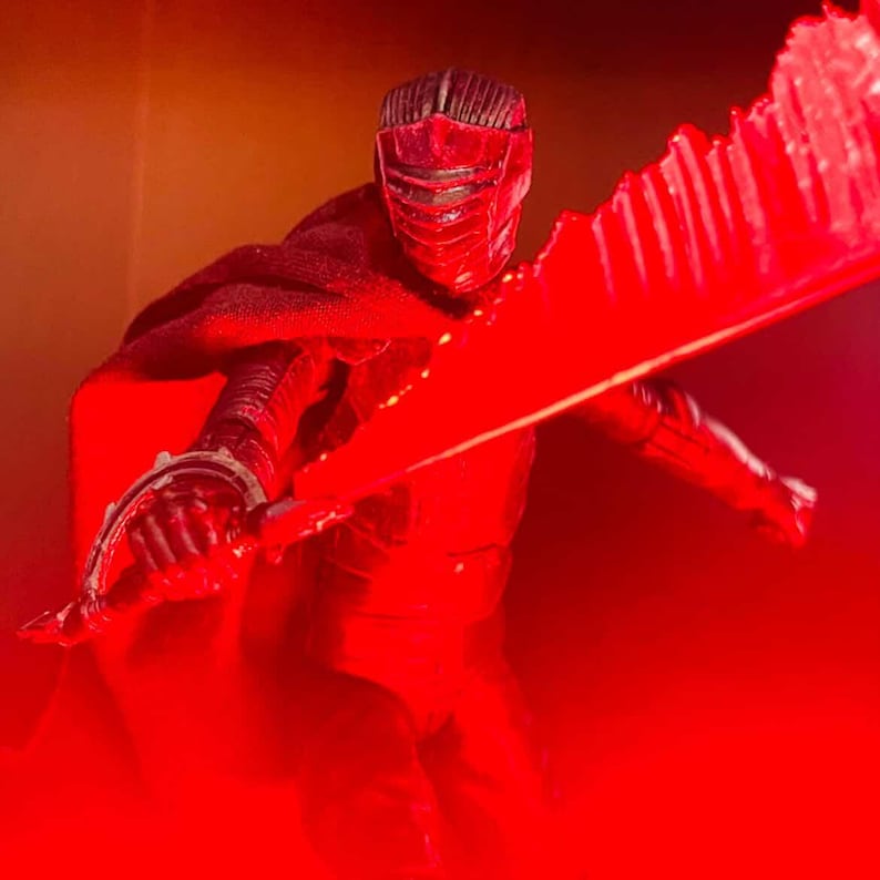 May include: A red action figure of a character in a black suit and helmet, holding a large red lightsaber. The figure is posed dynamically, with the lightsaber extended. The background is a gradient of orange and red.