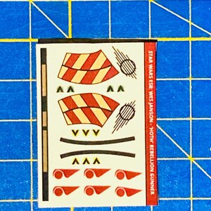 May include: A sheet of decals for a Star Wars model kit. The decals feature red and yellow stripes, black lines, and red circles. The text on the sheet reads "STAR WARS ESB: WES JANSON - 'HOTH' REBELLION GUNNER".