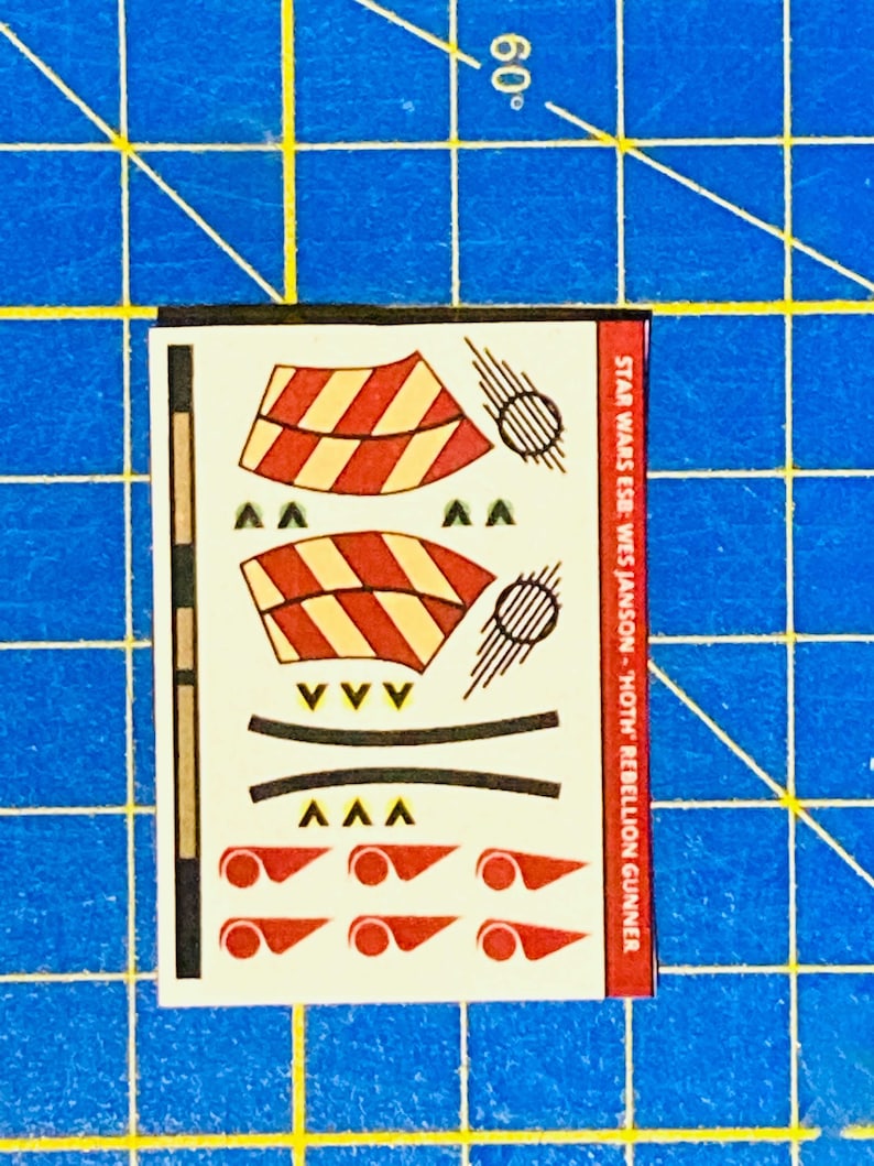May include: A sheet of decals for a Star Wars model kit. The decals feature red and yellow stripes, black lines, and red circles. The text on the sheet reads "STAR WARS ESB: WES JANSON - 'HOTH' REBELLION GUNNER".