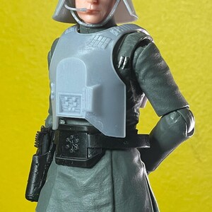 May include: A gray action figure of a character in a military uniform, complete with a helmet, chest armor, and a utility belt. The figure is holding a black pistol. The figure is standing in front of a yellow background.