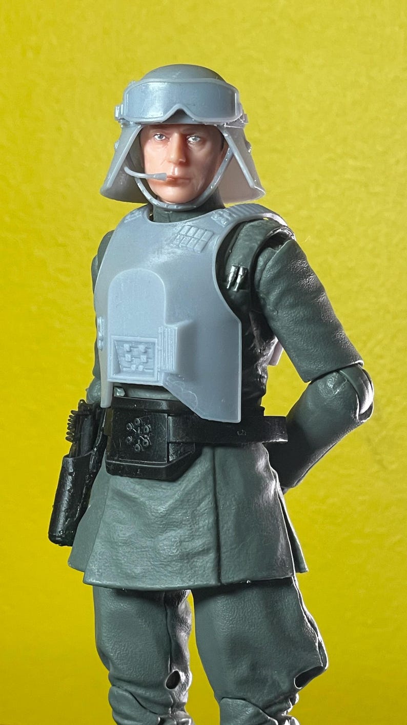 May include: A gray action figure of a character in a military uniform, complete with a helmet, chest armor, and a utility belt. The figure is holding a black pistol. The figure is standing in front of a yellow background.