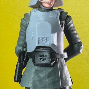 May include: A gray action figure of a character in a military uniform, complete with a helmet, chest armor, and a utility belt. The figure is holding a black weapon. The background is a solid yellow.