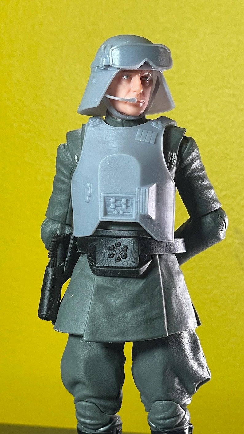 May include: A gray action figure of a character in a military uniform, complete with a helmet, chest armor, and a utility belt. The figure is holding a black weapon. The background is a solid yellow.